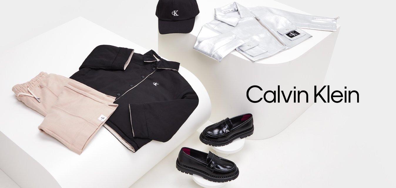 Giacca nera con bottoni, pantaloni jogger rosa pallido, giacca argentata con zip, cappellino nero e mocassini neri esposti su piattaforme geometriche bianche.