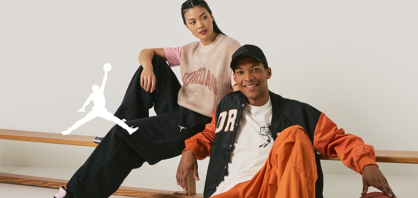 Due persone siedono casualmente su una panchina di legno. L'uomo indossa una giacca arancione e nera, una maglietta bianca, pantaloni arancioni e sneakers grigie. La donna indossa un maglione a maglia rosa e beige con pantaloni neri e sneakers nere.