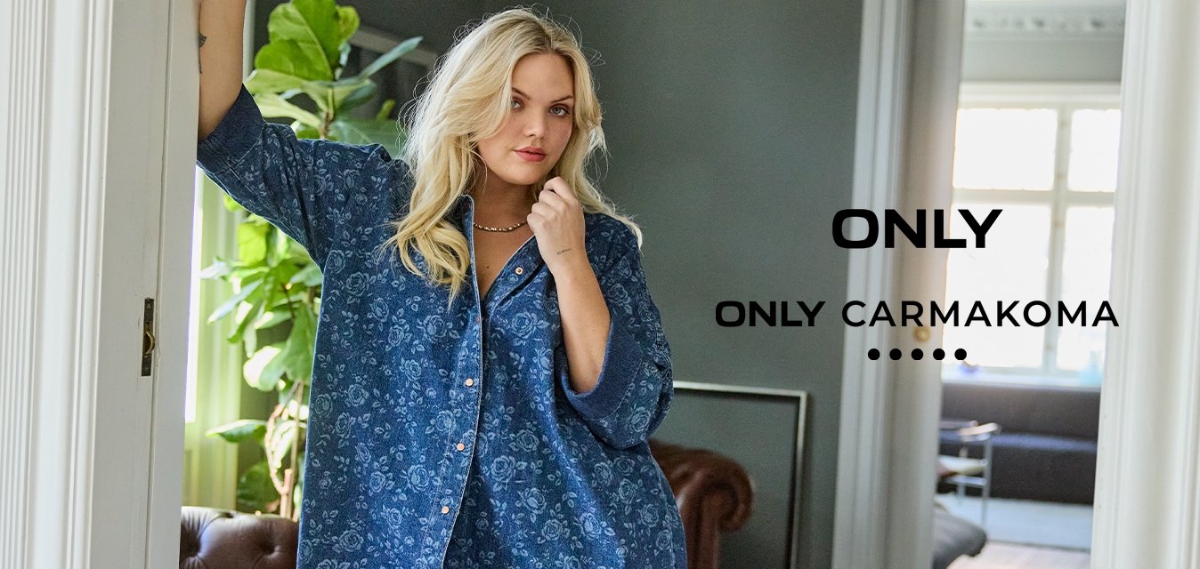 Donna con una maglietta floreale blu larga e jeans appoggiata a un telaio di porta in un soggiorno moderno e luminoso con piante e un divano in pelle.