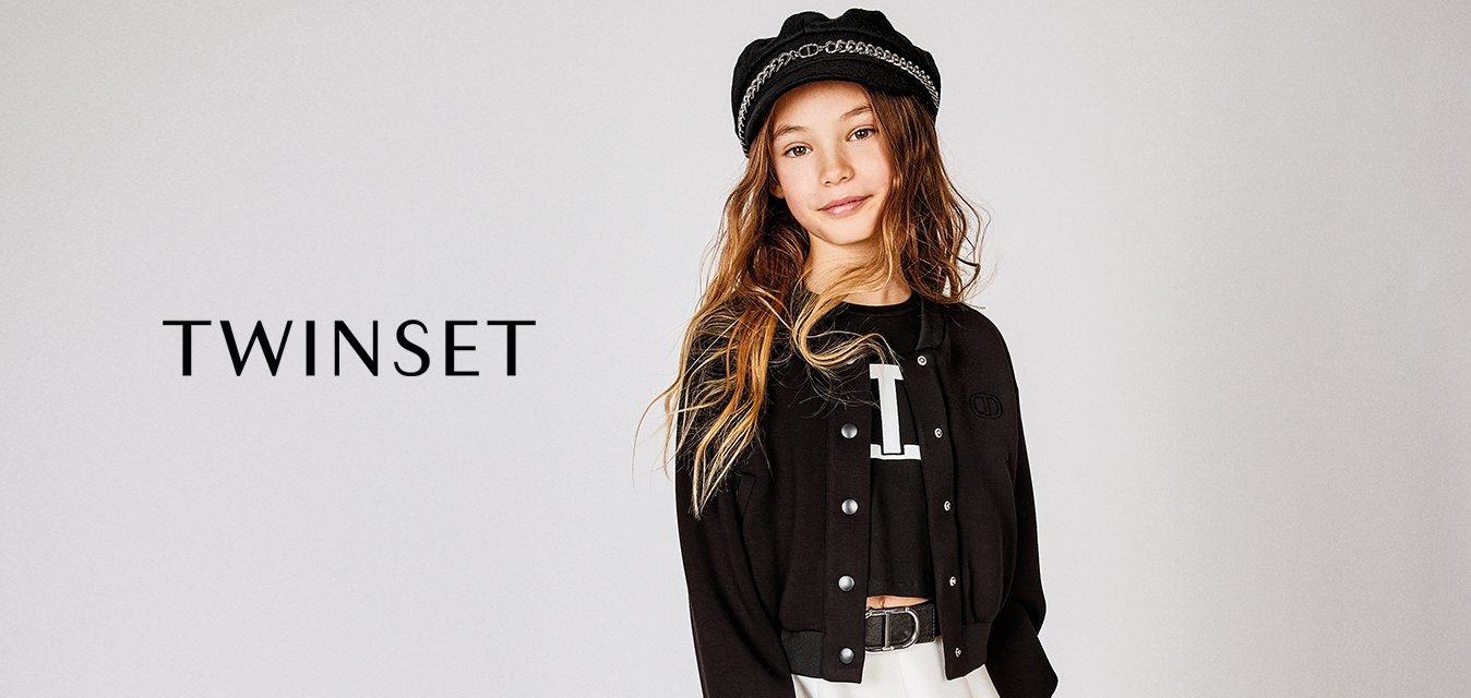 Giacca nera cropped con bottoni a pressione, indossata sopra una camicia nera con una lettera bianca. Gonna bianca svasata e un cappello nero con dettagli in catena.