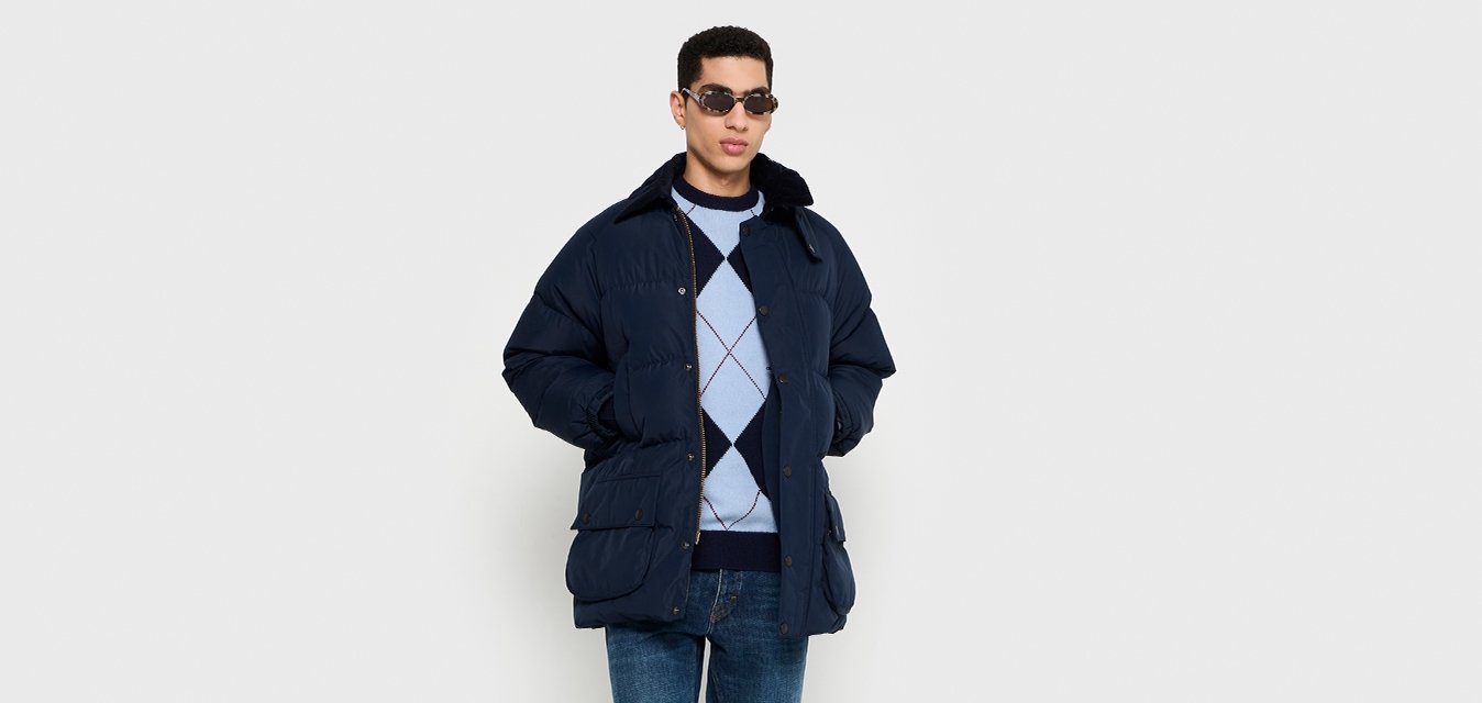 Giacca puffer blu navy con collo, maglione argyle blu scuro, jeans blu e scarpe nere; il modello indossa occhiali da sole.
