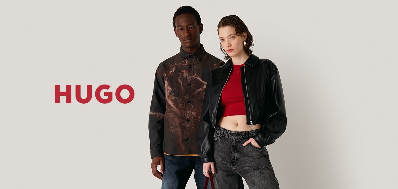 Due modelli si trovano di fronte a uno sfondo neutro. Uno indossa una camicia a fantasia marrone e jeans blu, l'altro una giacca cropped nera e pantaloni grigi.