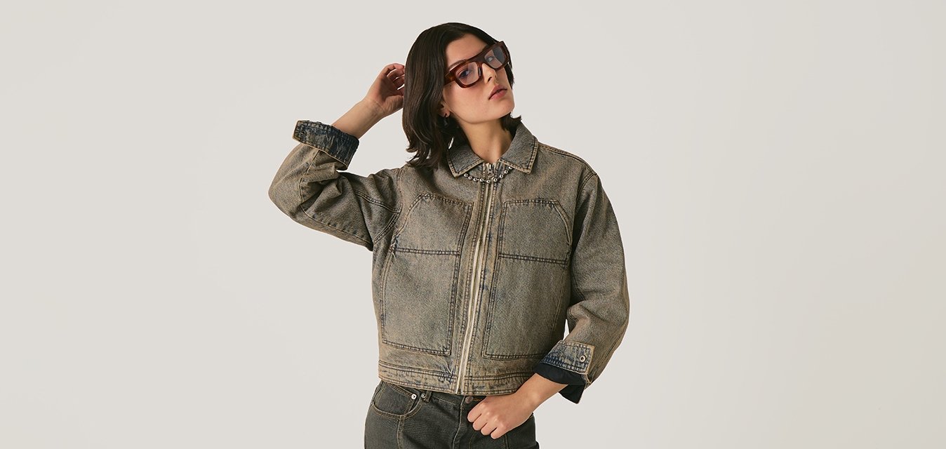 Donna che indossa un giubbotto di jeans oversize e una gonna abbinata, mentre sistema i capelli su uno sfondo neutro.