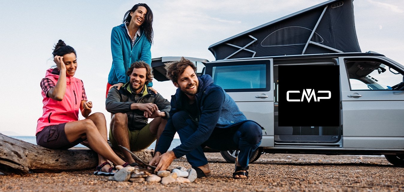 Quattro persone si sono riunite attorno a un piccolo falò su una superficie di ghiaia. Indossano abbigliamento outdoor colorato. Un furgone con il tetto sollevato è parcheggiato nelle vicinanze.