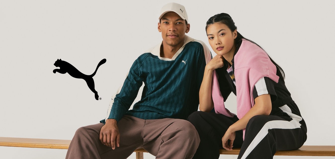 Due modelli siedono su una panchina di legno. L'uomo indossa una maglietta a righe verdi e pantaloni marroni, mentre la donna ha un tuta nera con dettagli rosa.