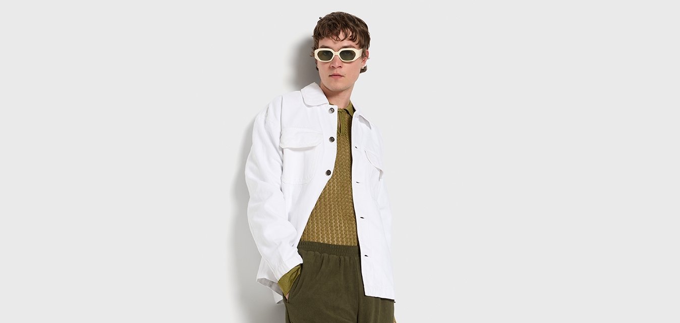 Giacca in denim bianca con due tasche frontali, abbinata a una maglia verde oliva texture e a pantaloni verdi scuri. Modella con occhiali da sole chiari.