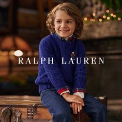 Bambino sorridente con capelli ricci, vestito con un maglione azzurro a trecce e jeans, seduto su un antico baule di legno in una stanza accogliente con camino in pietra.