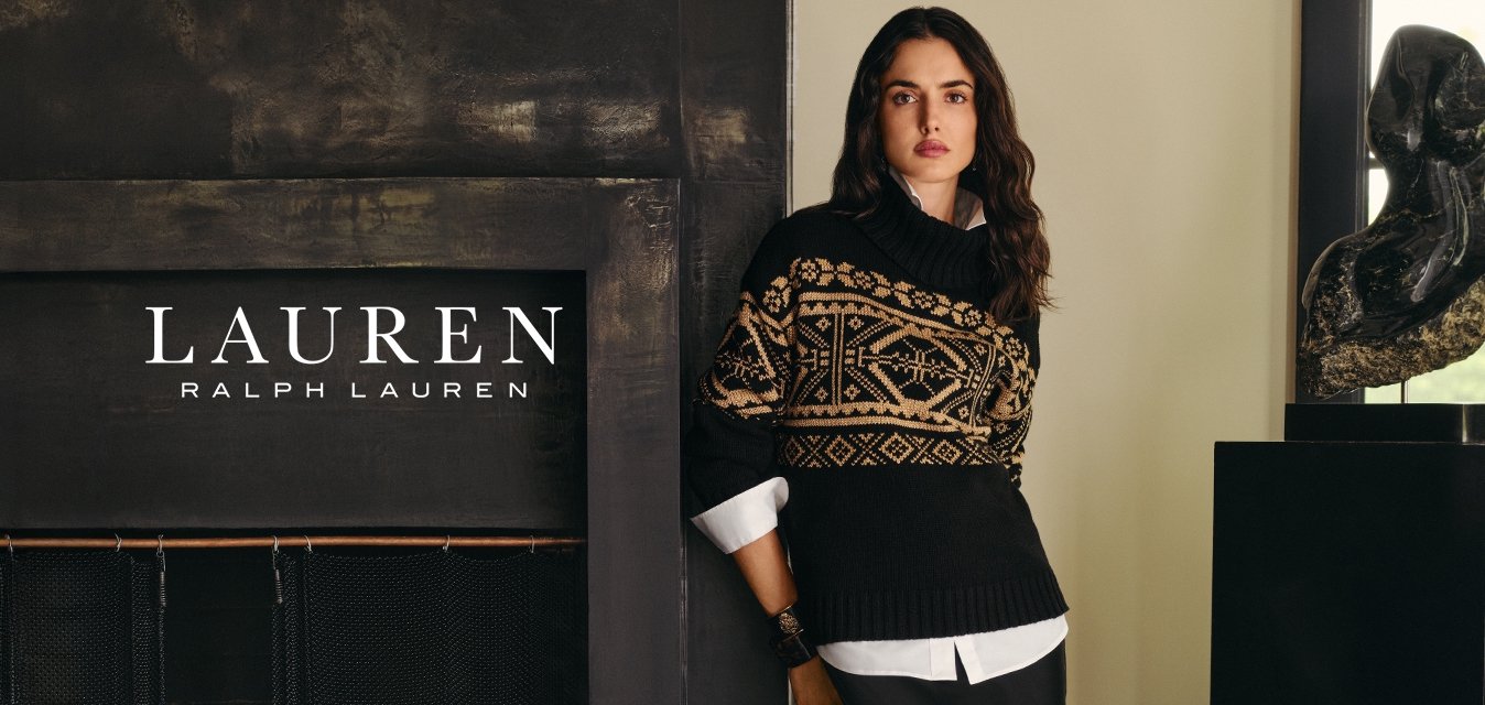 Donna in nero, maglione con motivo beige e lunga gonna nera si trova vicino a un caminetto scuro e a una scultura astratta su un piedistallo vicino alla finestra.