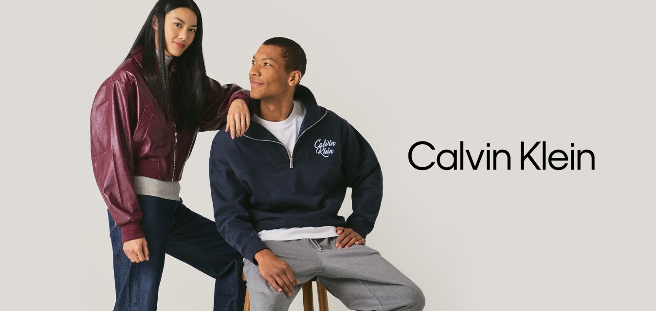 Giovane donna con giacca bordeaux e jeans sta accanto a un uomo in felpa blu navy e pantaloni grigi seduto su uno sgabello contro uno sfondo semplice.