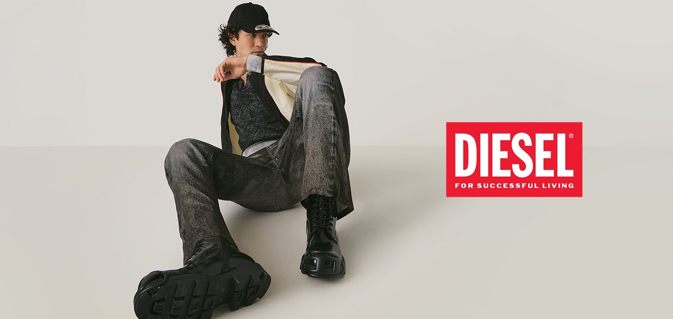 Una persona con un berretto nero, una giacca bianca e nera, pantaloni grigi e stivali neri è seduta sul pavimento contro uno sfondo chiaro e semplice.