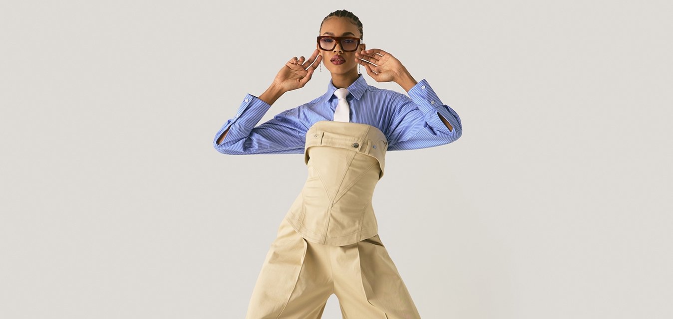 Modella in posa con pantaloni beige a gamba larga e corsetto sopra una camicia a righe blu con cravatta bianca, accessoriata con occhiali e orecchini.