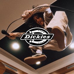 Cantante con tatuaggi salta in uno studio musicale tenendo un microfono, un batterista suona dietro un set di batteria, attrezzature moderne per studio sono visibili.