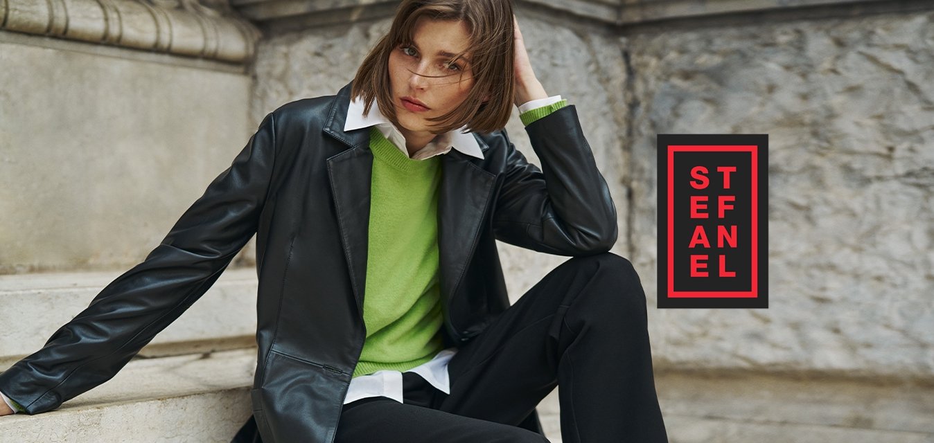 Blazer di pelle nera sopra un maglione verde, camicia bianca e pantaloni neri a gamba larga. Il modello è seduto su scale di marmo con uno sfondo texturizzato.