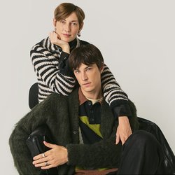 Due giovani adulti, uno seduto e l'altro in piedi dietro con un braccio sulla spalla, entrambi indossano maglioni a trama e a righe su uno sfondo semplice.