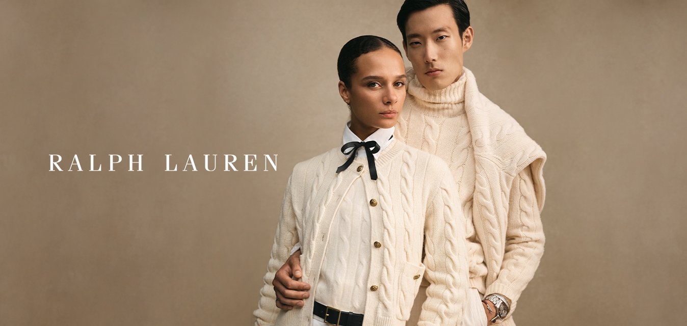 Due modelli che indossano maglioni a treccia color crema posano vicino su uno sfondo beige, uno con un papillon e l'altro con un orologio.