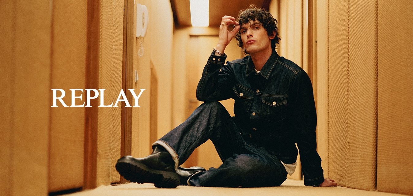 Giovane in giacca di jeans e pantaloni di denim è seduto sul pavimento di moquette di un stretto corridoio beige, toccandosi i capelli con una mano.