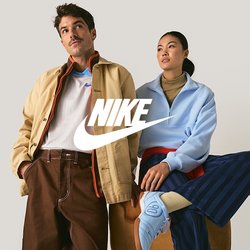 Uomo con giacca beige e pantaloni marroni sta accanto a una donna con pullover azzurro chiaro e pantaloni a righe blu scuro seduta su una panchina.