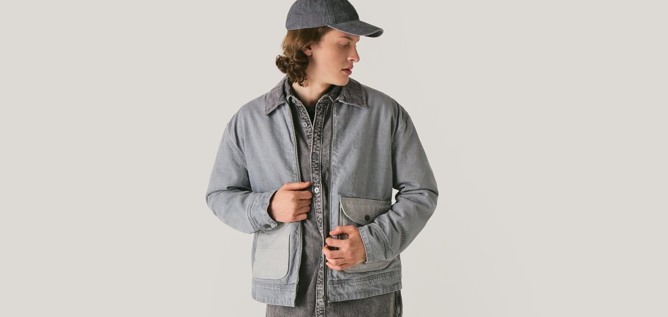 Giovane che indossa una giacca di denim grigia, un berretto abbinato e una maglia scura, che guarda in basso a sinistra su uno sfondo chiaro e semplice.