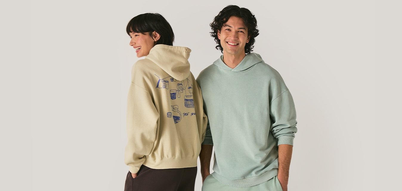 Uomo e donna sorridenti che indossano felpe pastello e pantaloni comodi, in piedi fianco a fianco contro uno sfondo chiaro e semplice.