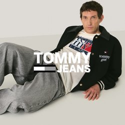 Uomo sdraiato sul pavimento che indossa una giacca nera, una maglietta bianca con grafica, jeans grigi e scarpe da ginnastica bianche, con uno sfondo beige neutro.