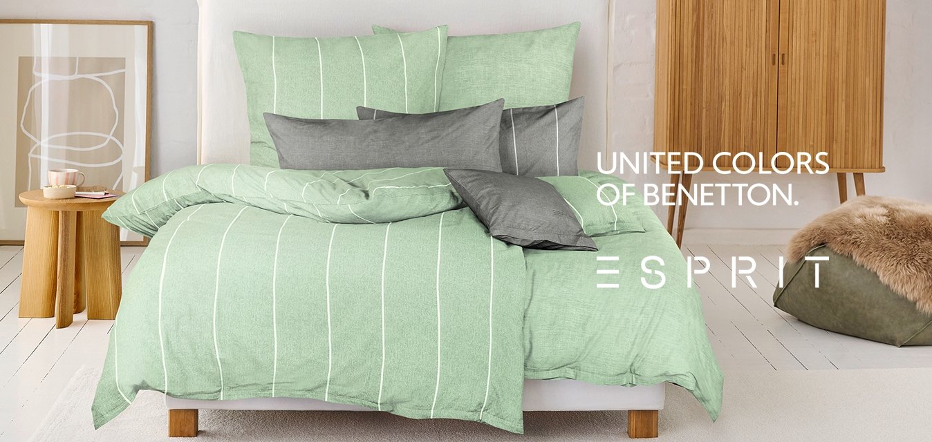 Set di biancheria da letto a righe verdi su un letto con vari cuscini grigi. Mobili in legno chiaro e un pouf dalle texture morbide in una stanza minimalista.