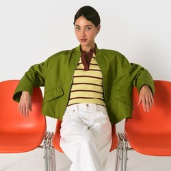 Donna seduta con le gambe incrociate su una sedia arancione a metà, con le braccia appoggiate su sedie vuote adiacenti, indossa una giacca verde e scarpe rosse.