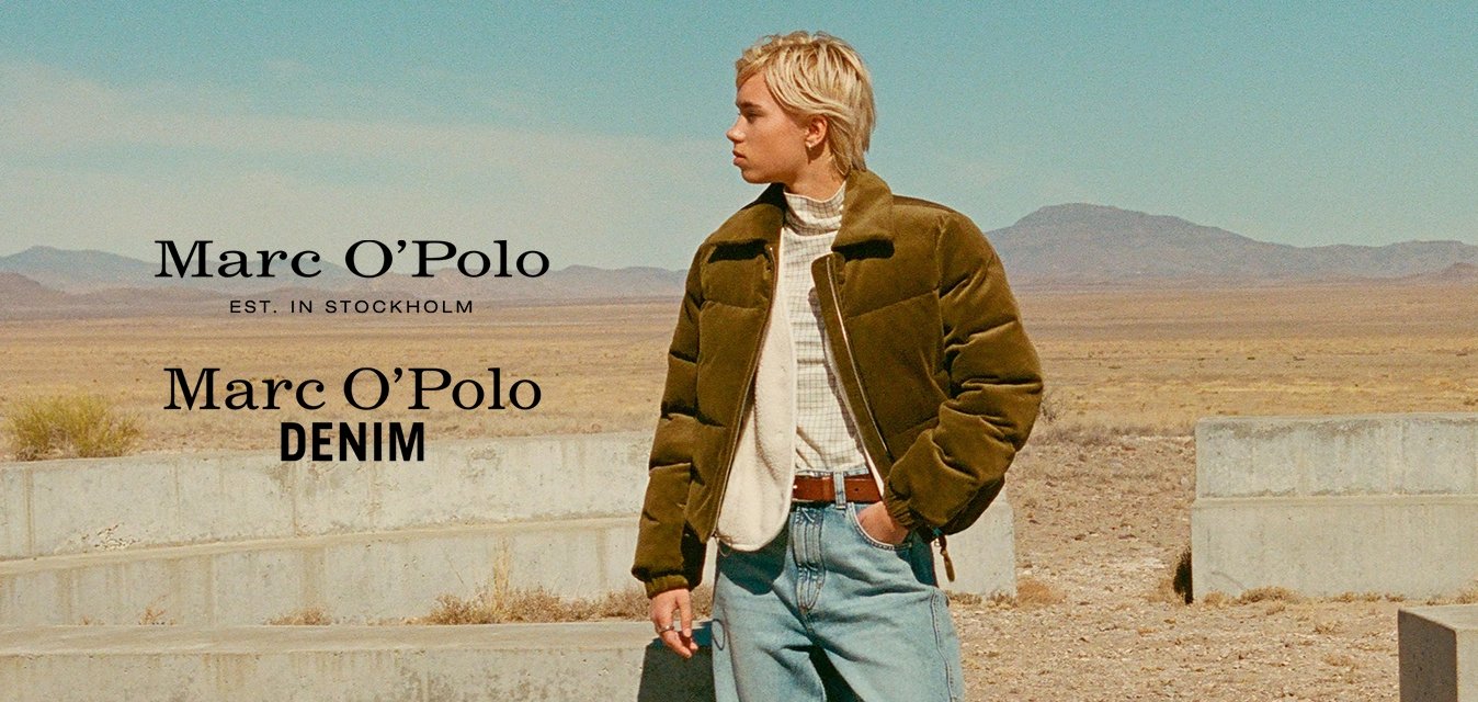 Giacca puffer verde oliva sopra un maglione crema, jeans azzurri chiari, stivaletti beige; in piedi vicino a strutture in cemento grigio in un paesaggio desertico.