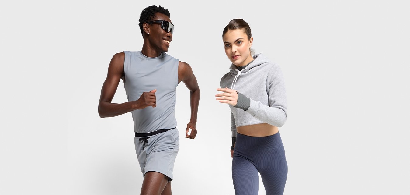 Due persone che corrono indoors indossando abbigliamento sportivo; una in pantaloni corti grigi e top senza maniche, l'altra in leggings blu navy e felpa grigia corta.