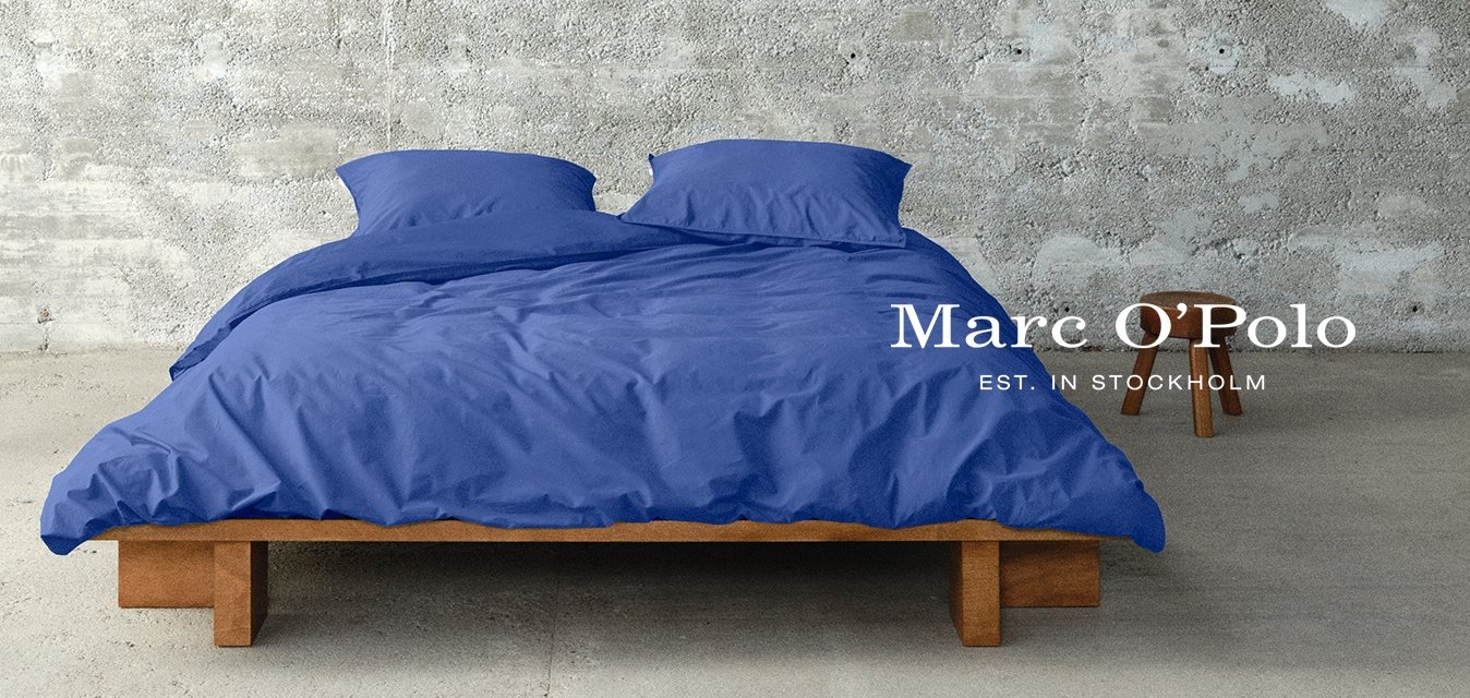 Struttura del letto in legno con un piumone blu e cuscini coordinati. Parete e pavimento in cemento, con un piccolo sgabello di legno nelle vicinanze. Design minimalista.