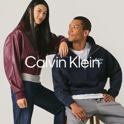 Giovane donna con giacca bordeaux e jeans sta accanto a un uomo in felpa blu navy e pantaloni grigi seduto su uno sgabello contro uno sfondo semplice.