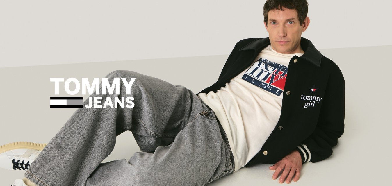 Uomo sdraiato sul pavimento che indossa una giacca nera, una maglietta bianca con grafica, jeans grigi e scarpe da ginnastica bianche, con uno sfondo beige neutro.