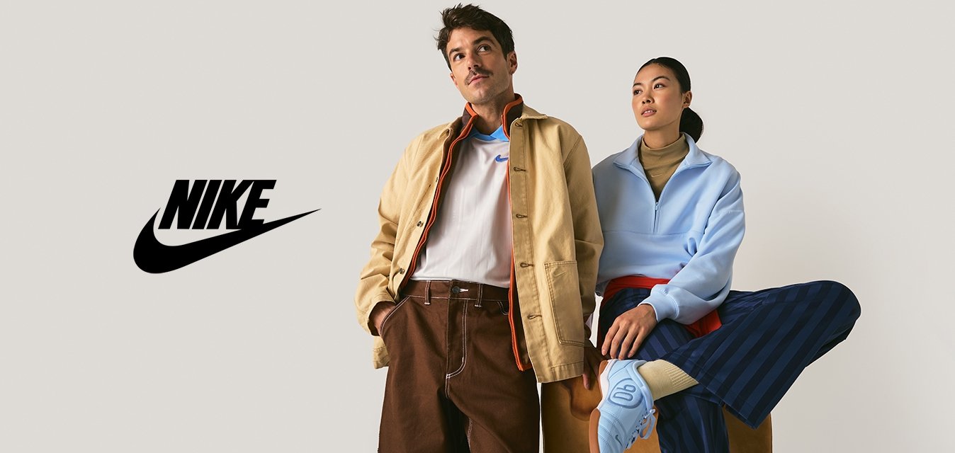 Uomo con giacca beige e pantaloni marroni sta accanto a una donna con pullover azzurro chiaro e pantaloni a righe blu scuro seduta su una panchina.