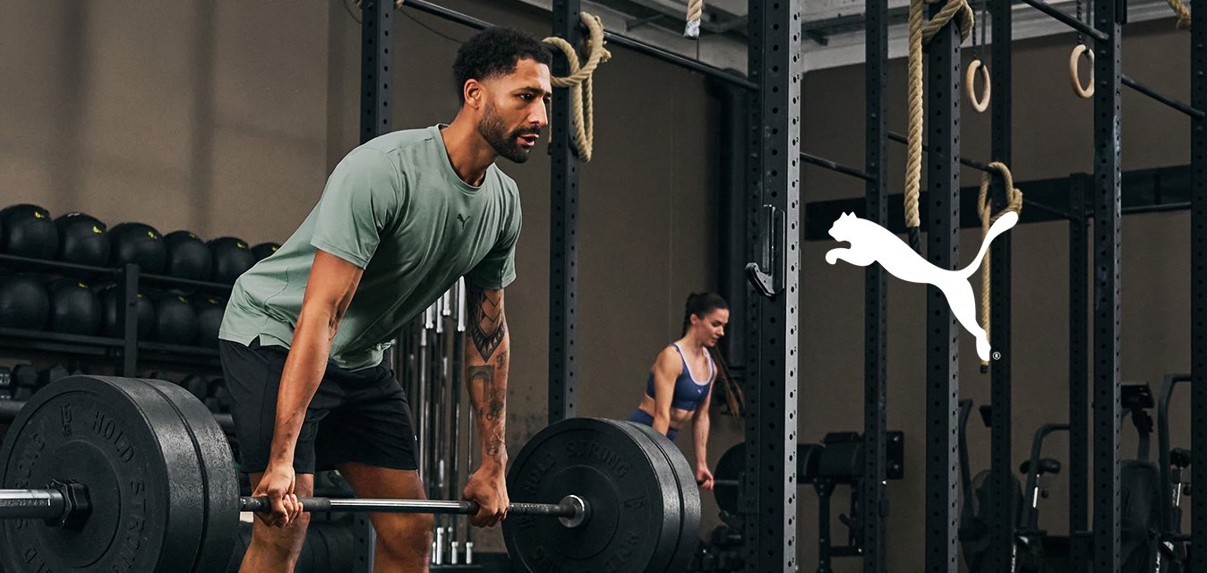 Cinque persone che si allenano in una palestra; uno solleva un bilanciere, gli altri utilizzano palle mediche o attrezzature da palestra in un'area di allenamento ampia e ben illuminata.