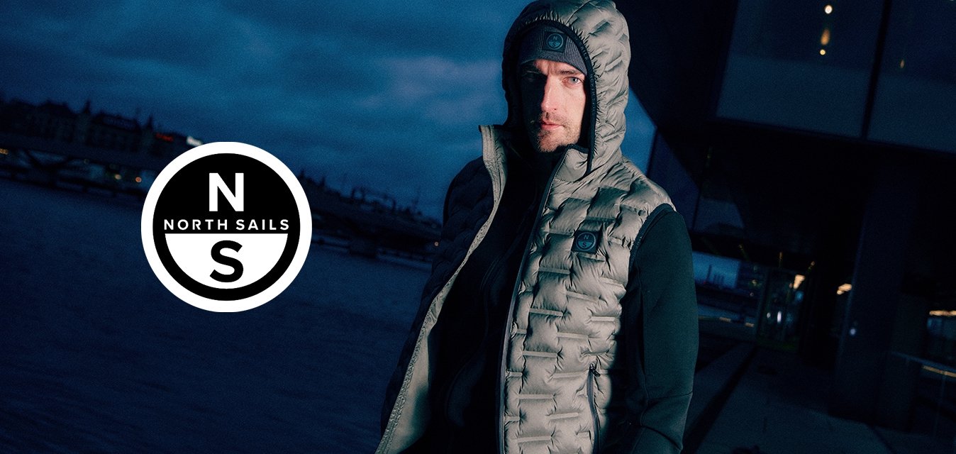 Gilet imbottito grigio con cappuccio, caratterizzato da un design trapuntato e una toppa con logo, indossato sopra una maglia nera a maniche lunghe, sullo sfondo di un lungomare.