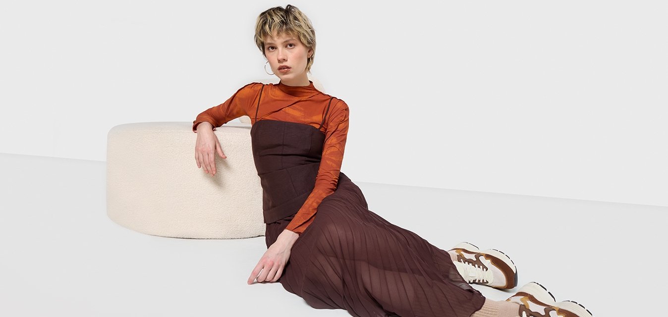 Gonna a pieghe lunga marrone con un top scuro attillato, abbinata a sneaker marroni e bianche. Vicino, un morbido pouf cilindrico bianco.