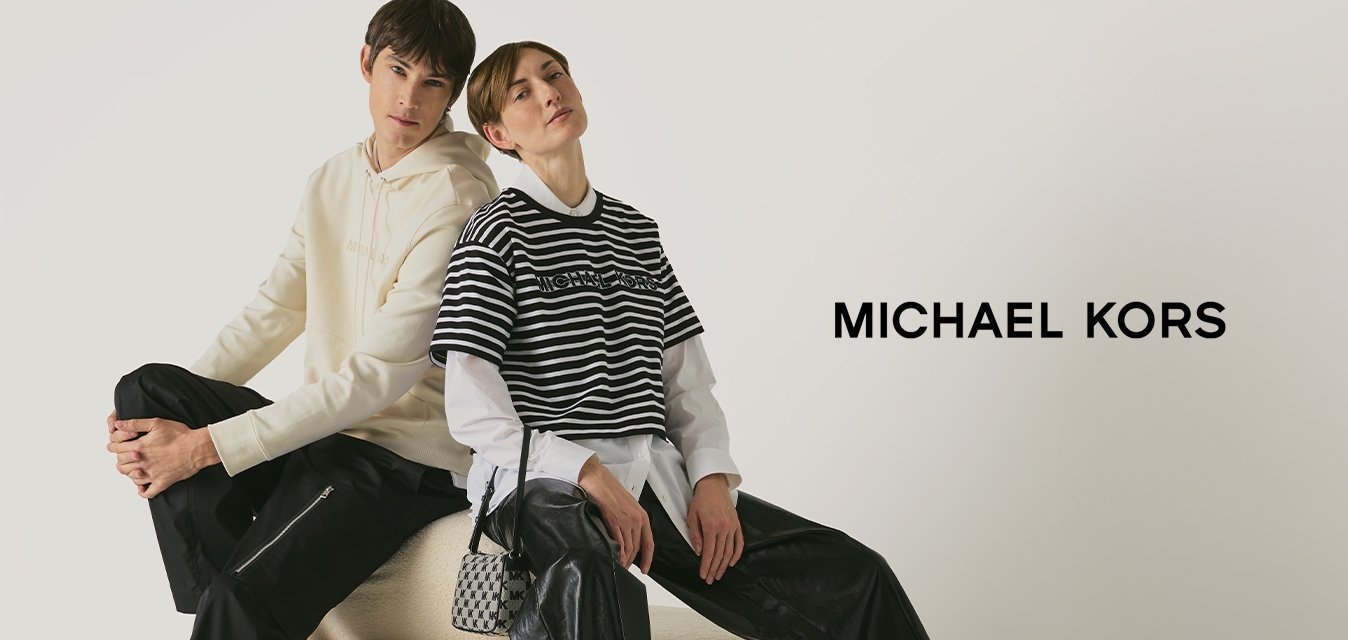 Due giovani seduti schiena contro schiena su un ottomano rotondo beige, vestiti con outfit casual chic, su uno sfondo bianco.