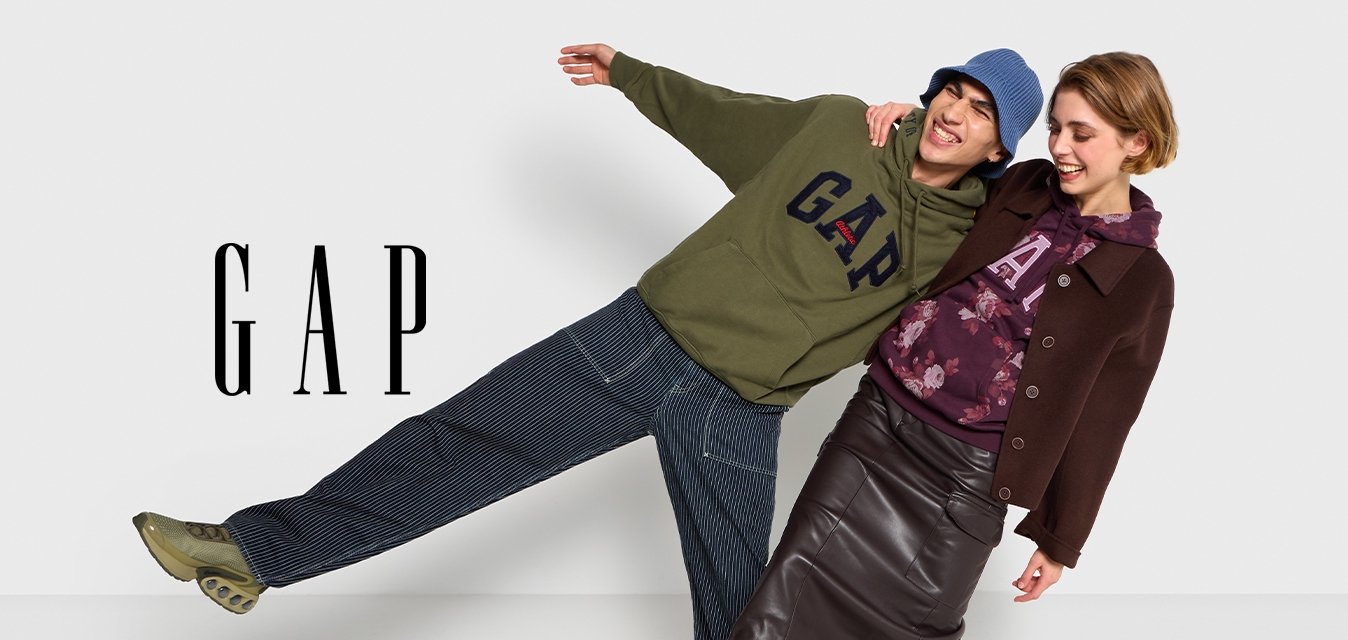 Due modelli si appoggiano in modo giocoso a un muro, indossando abiti casual. Uno indossa una felpa verde oliva e pantaloni larghi, l'altro un cappotto marrone scuro e una gonna.