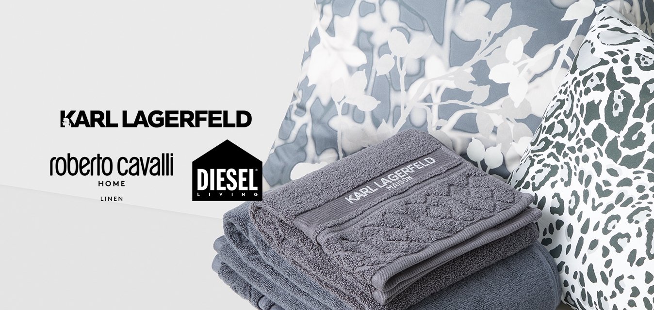 Pila di asciugamani grigi Karl Lagerfeld su una superficie bianca con cuscini a stampa floreale e leopardata sullo sfondo.