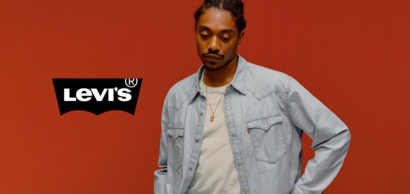 Uomo con i capelli intrecciati che indossa una camicia di jeans chiara e una collana, in piedi con una mano sui fianchi, contro uno sfondo arancione uniforme.