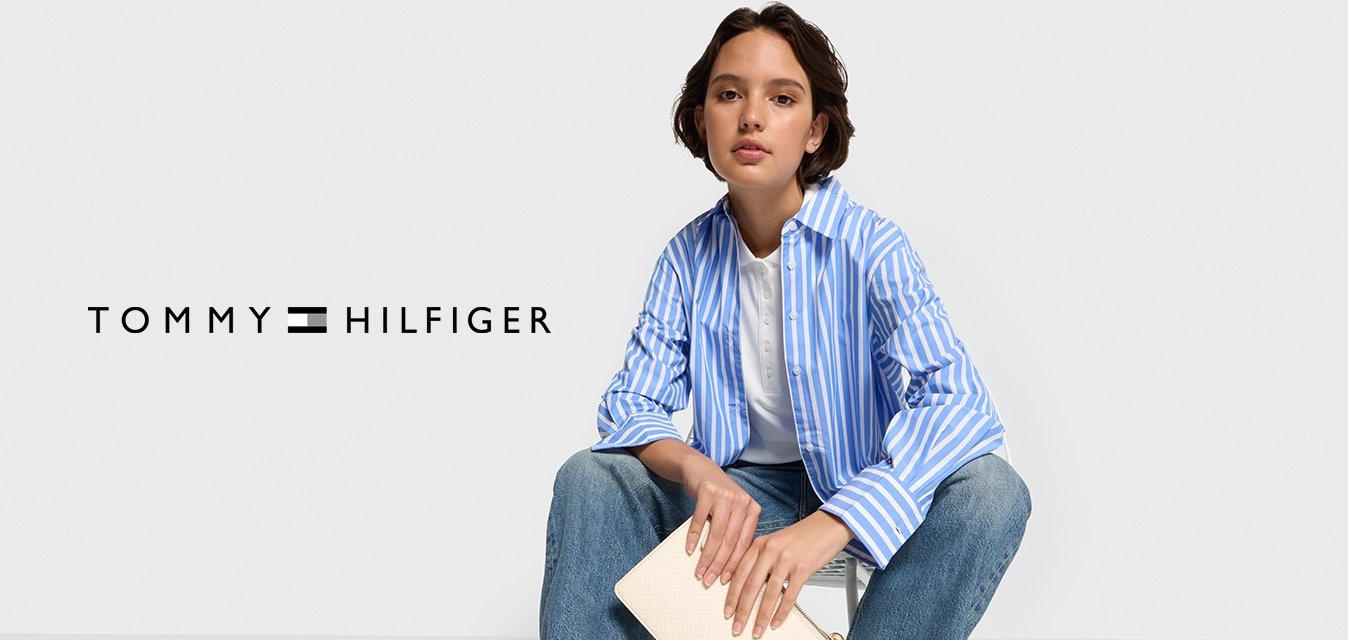 Camicia a maniche lunghe a righe blu e bianche, t-shirt bianca, jeans azzurri chiari e sneakers bianche. Tieni in mano una pochette color crema con un logo.