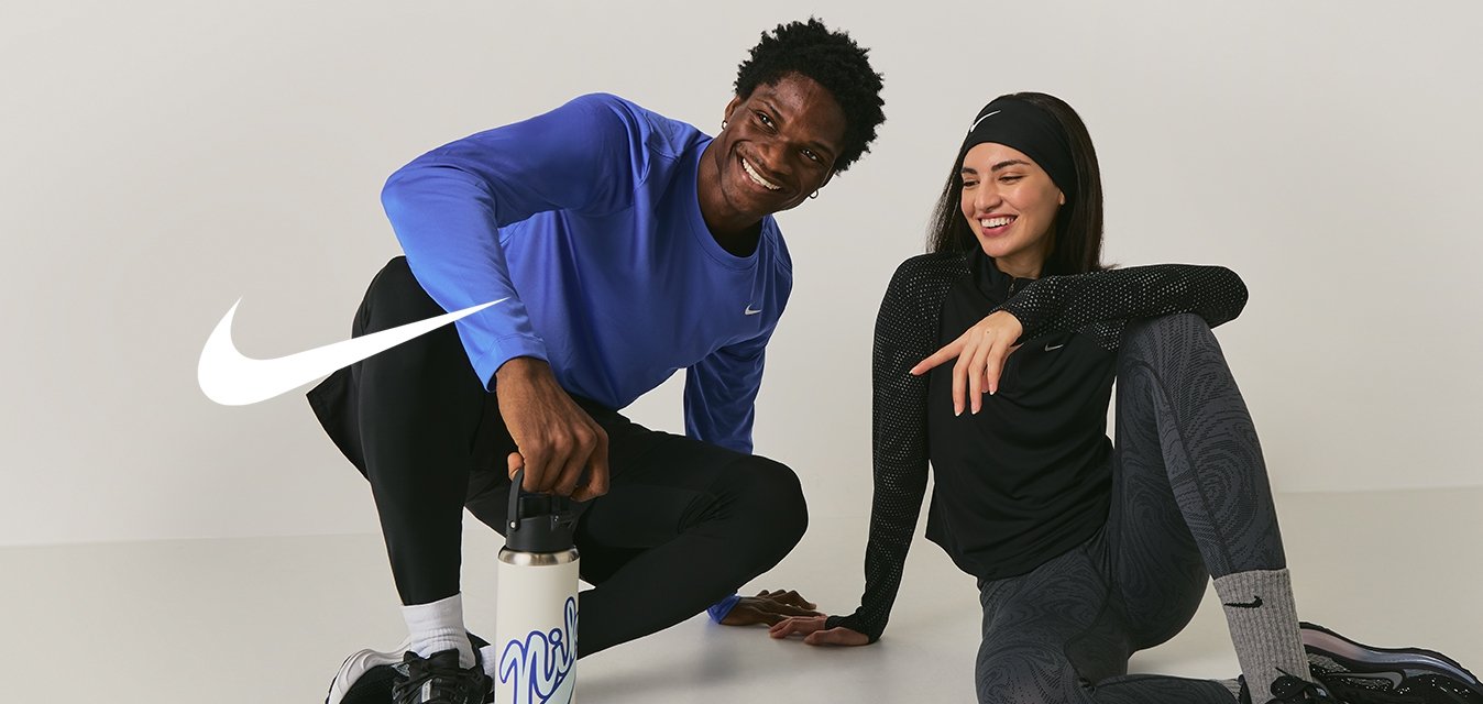 Due persone in abbigliamento sportivo sedute a terra, sorridenti; una tiene una bottiglia d'acqua, entrambe sembrano rilassate e amichevoli.