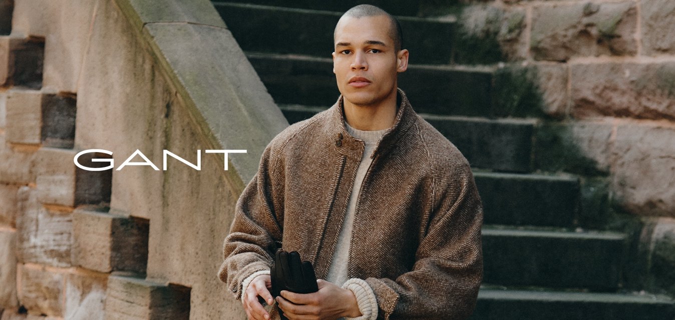 Giacca bomber marrone strutturata sopra un maglione in maglia crema, jeans scuri e scarpe nere. In piedi su gradini di pietra con un sentiero visibile.