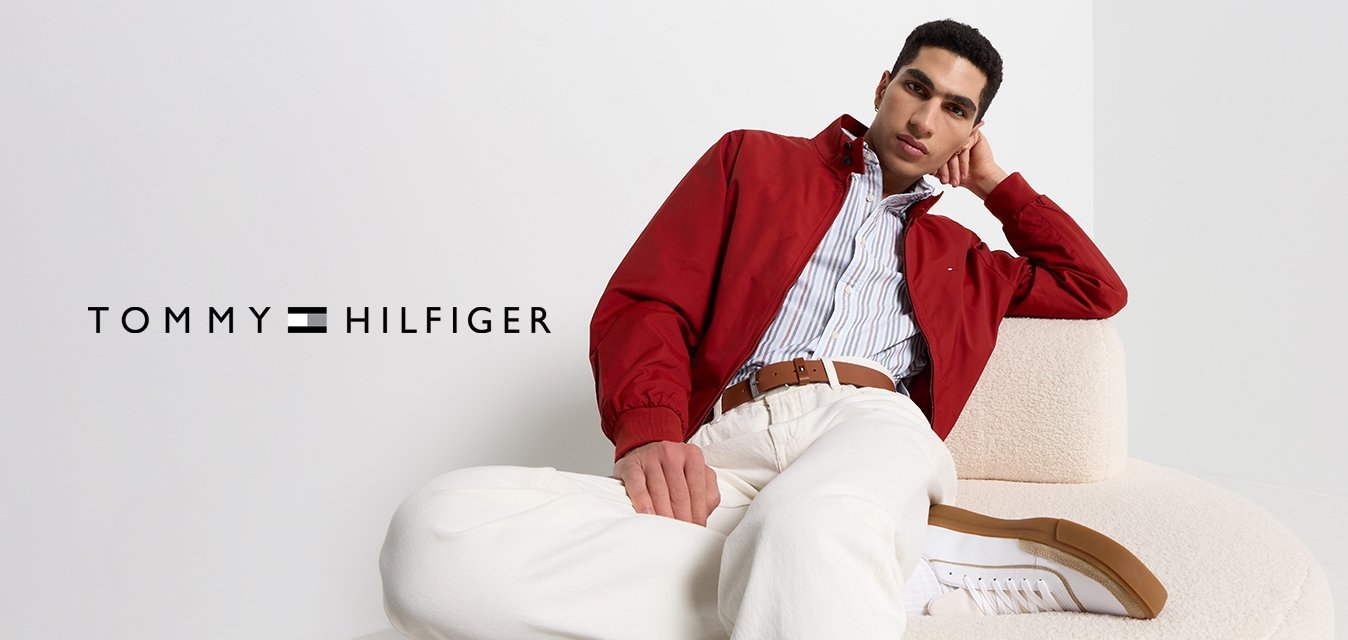 Giovane uomo in giacca rossa, camicia a righe, pantaloni bianchi e sneakers che si rilassa su un divano beige curvo contro uno sfondo bianco semplice.