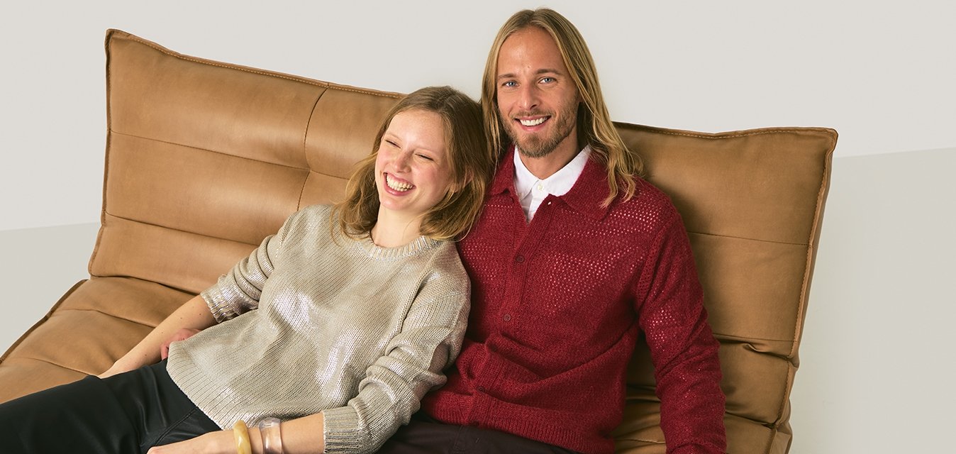 Due persone sorridenti, sedute vicine su un divano con cuscini color cuoio; una indossa un maglione argento metallico, l'altra un cardigan rosso lavorato a maglia.