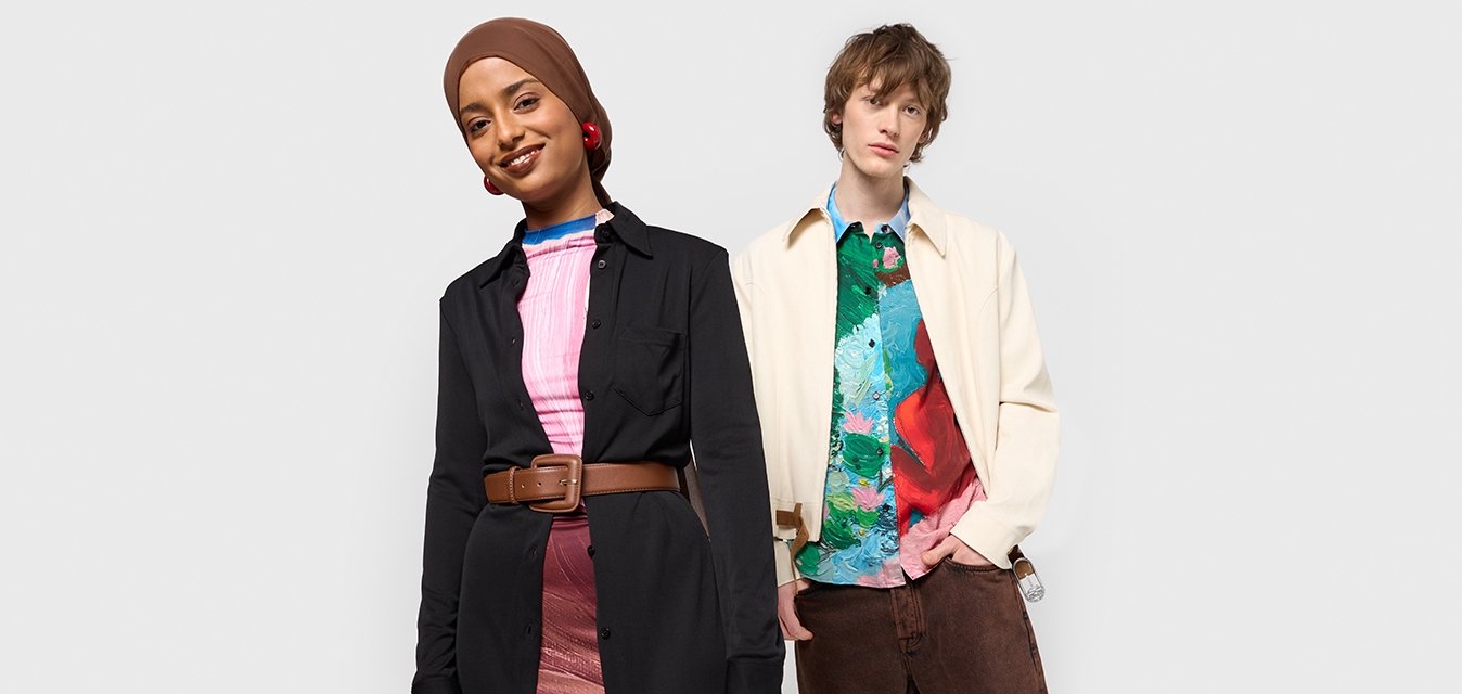 Donna in un abito nero con cintura marrone e gonna a fantasia, che indossa un foulard marrone; uomo con camicia colorata, giacca crema e pantaloni marroni, in piedi fianco a fianco.