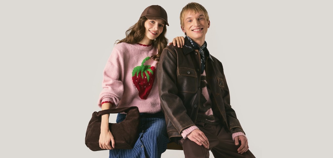 Gio giovane in un maglione rosa a fragola, gonna blu e berretto marrone si siede accanto a un uomo sorridente in giacca di pelle marrone e sciarpa su uno sfondo beige.