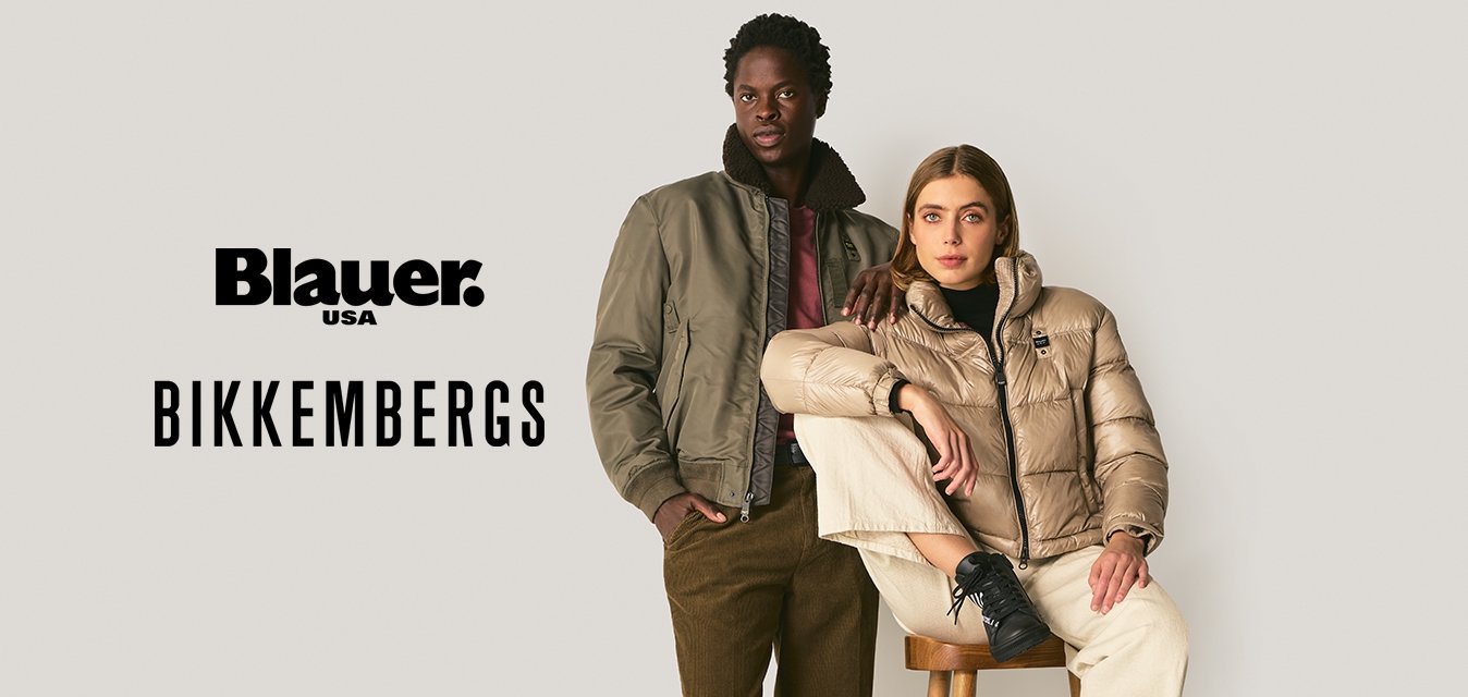 Due adulti indossano giacche calde: uno in piedi con un bomber oliva e pantaloni marroni, l'altro seduto su uno sgabello con un piumino beige e pantaloni crema.